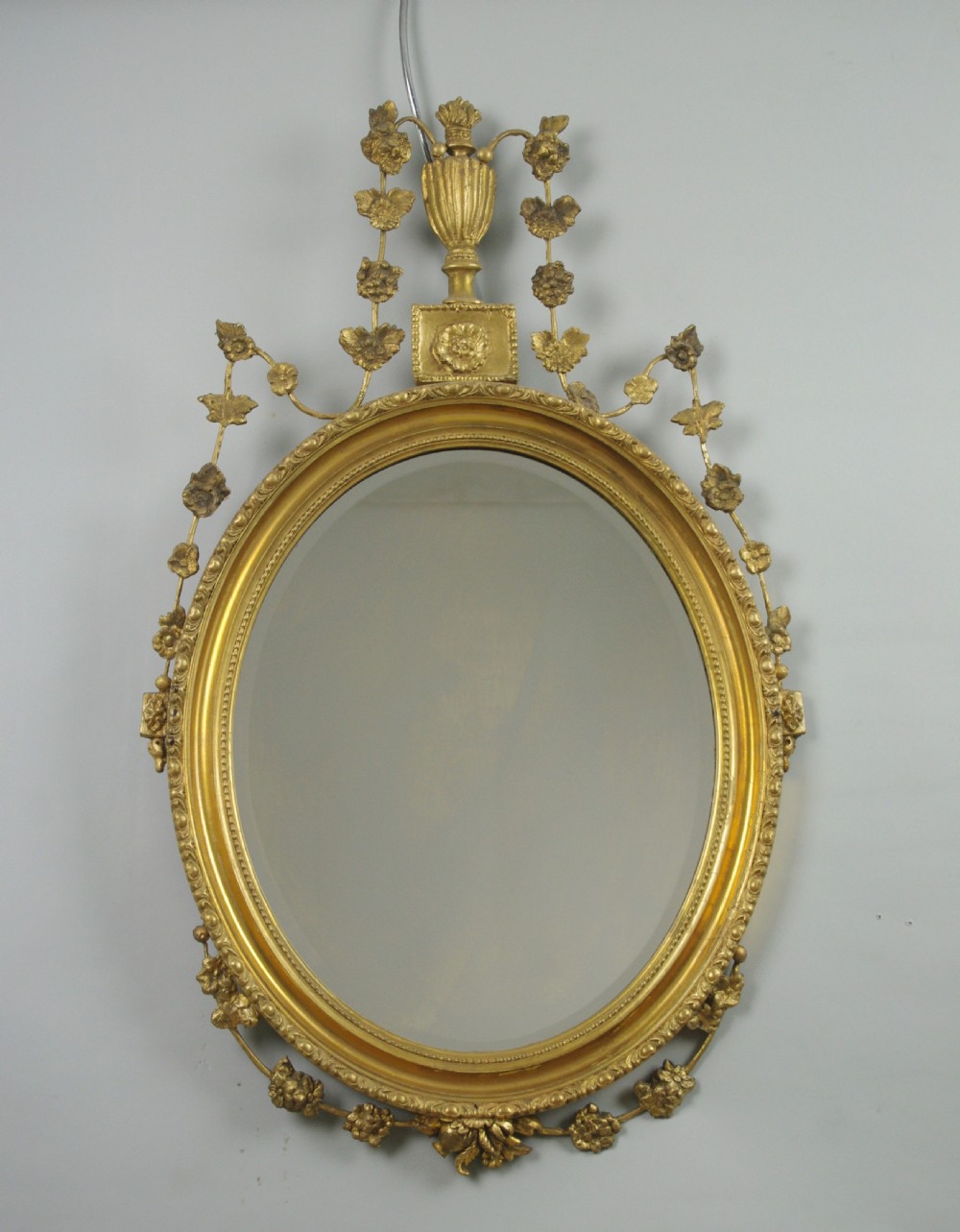A Victorian oval wall mirror featuring a gilded frame embellished with beaded moulding, an urn finial, and delicate leaf garlands.
