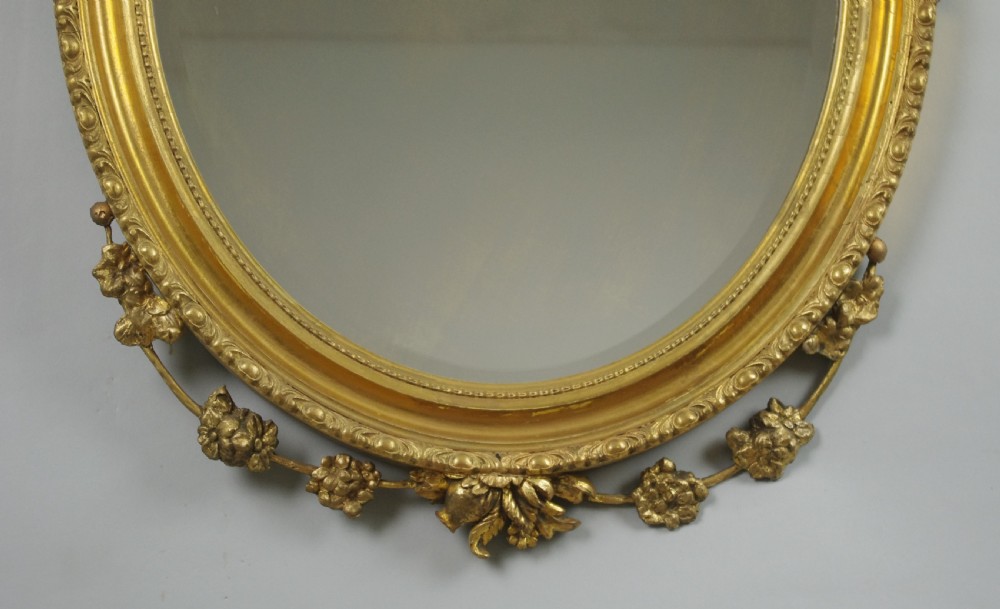 A Victorian oval wall mirror featuring a gilded frame embellished with beaded moulding, an urn finial, and delicate leaf garlands. View VI