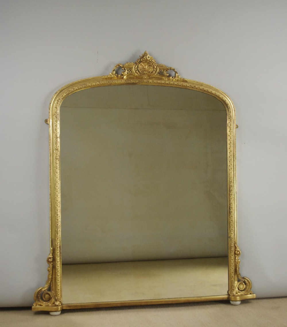 An arched Victorian overmantel mirror featuring a gilded frame with floral mouldings, scrolled side feet, and an ornate top crest.