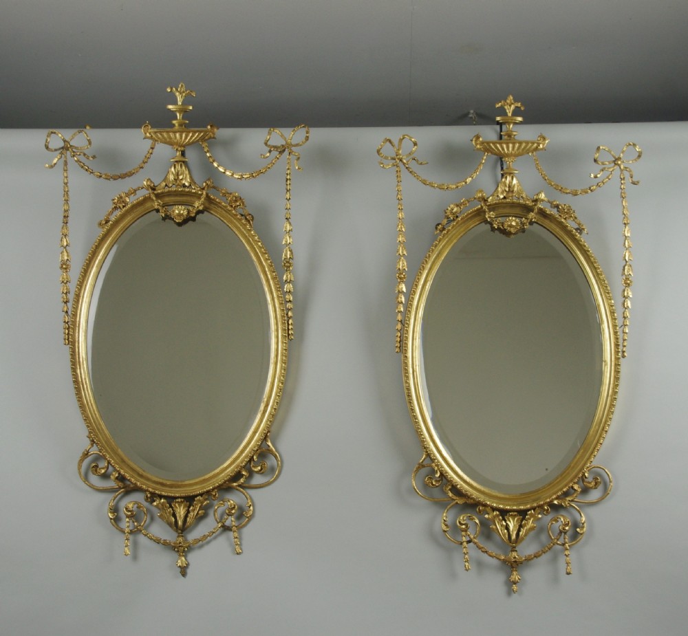 A pair of Victorian giltwood oval wall mirrors topped with urn crestings and ribbon bow swags.