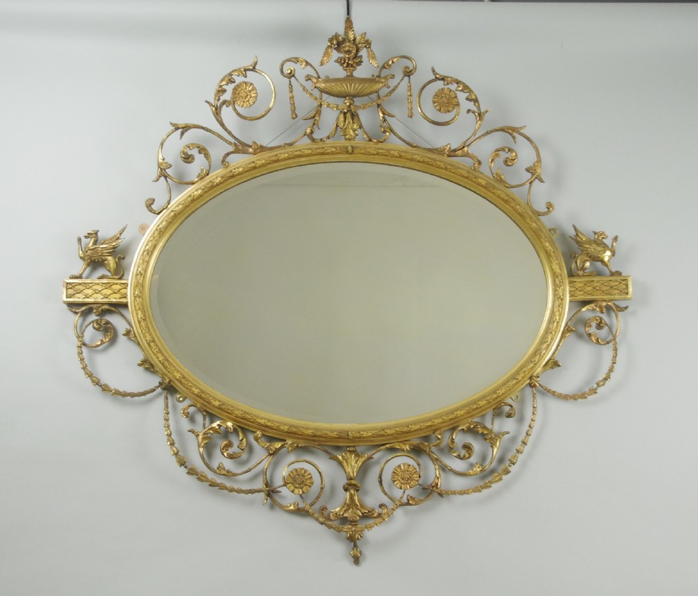 A Victorian giltwood oval wall mirror in the Adam style featuring scrollwork, urn cresting, griffin accents, and floral garlands.