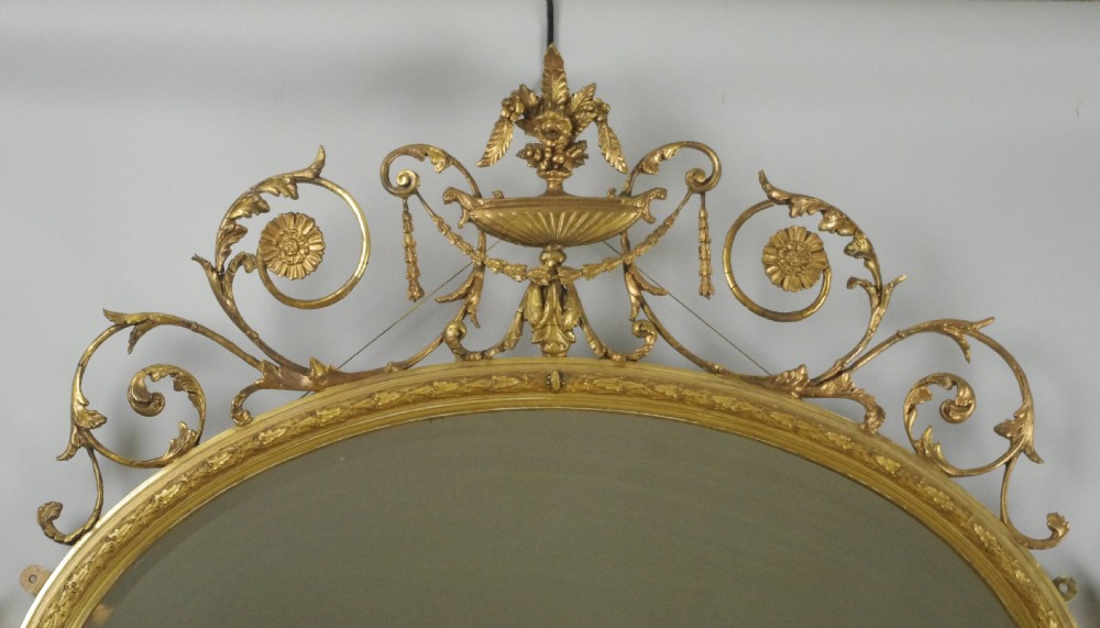 A Victorian giltwood oval wall mirror in the Adam style featuring scrollwork, urn cresting, griffin accents, and floral garlands. View V