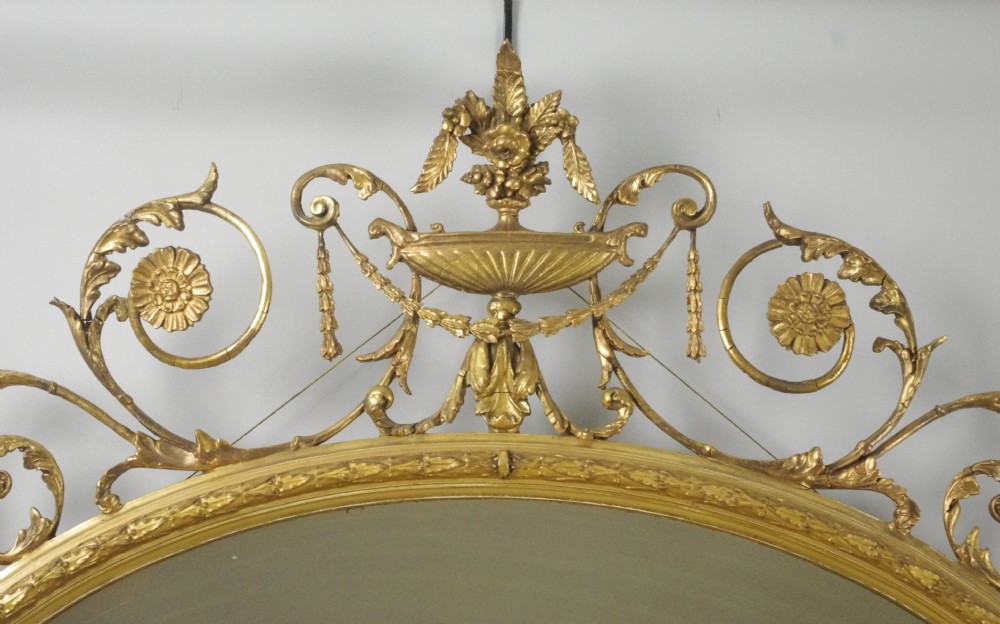 A Victorian giltwood oval wall mirror in the Adam style featuring scrollwork, urn cresting, griffin accents, and floral garlands. View VI
