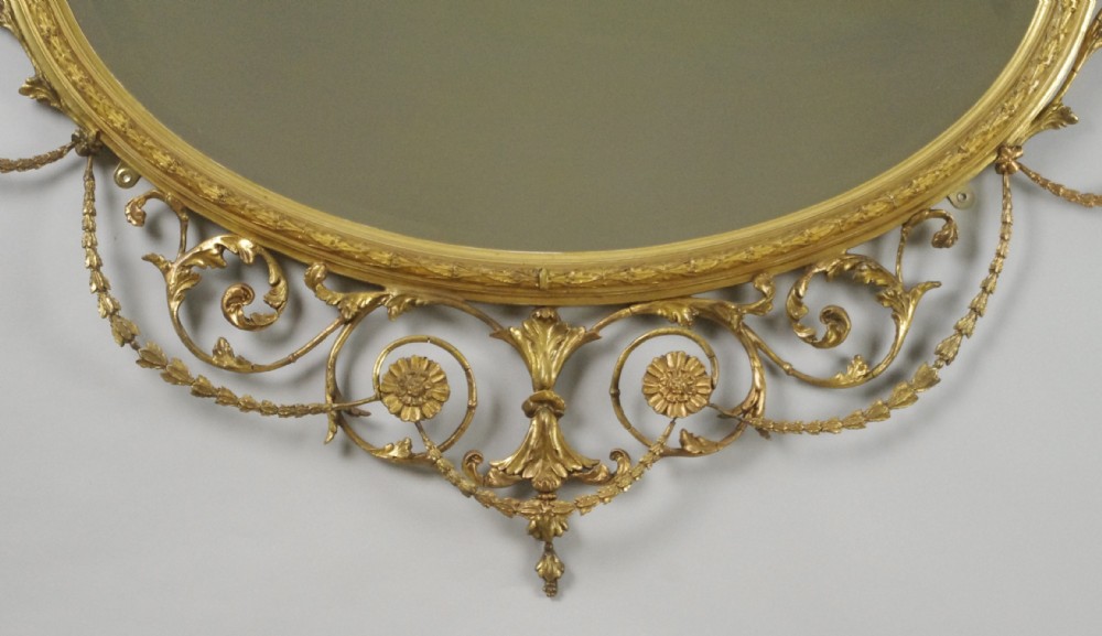 A Victorian giltwood oval wall mirror in the Adam style featuring scrollwork, urn cresting, griffin accents, and floral garlands. View VII