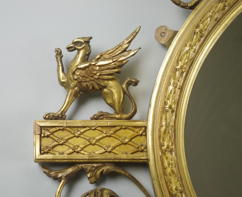 A Victorian giltwood oval wall mirror in the Adam style featuring scrollwork, urn cresting, griffin accents, and floral garlands. View IX