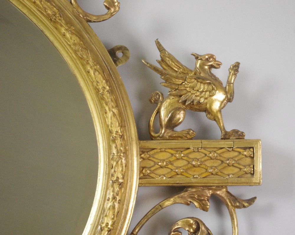 A Victorian giltwood oval wall mirror in the Adam style featuring scrollwork, urn cresting, griffin accents, and floral garlands. View X