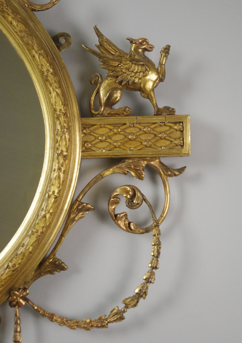 A Victorian giltwood oval wall mirror in the Adam style featuring scrollwork, urn cresting, griffin accents, and floral garlands. View XII