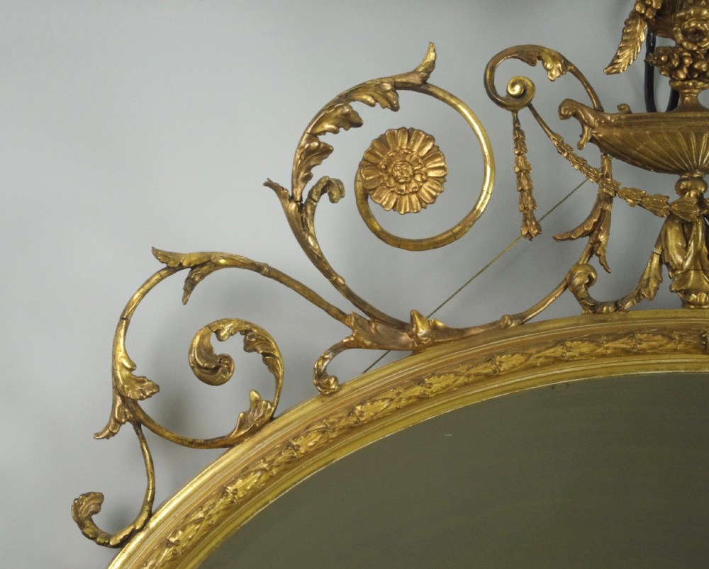 A Victorian giltwood oval wall mirror in the Adam style featuring scrollwork, urn cresting, griffin accents, and floral garlands. View XIII