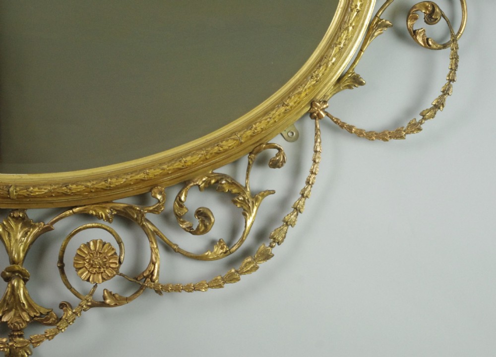 A Victorian giltwood oval wall mirror in the Adam style featuring scrollwork, urn cresting, griffin accents, and floral garlands. View XIV