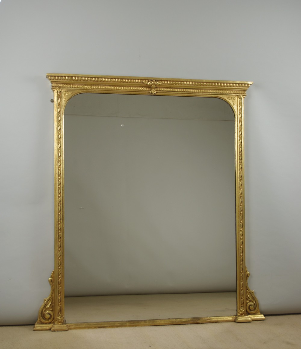 A large Victorian gilt overmantel mirror featuring a rectangular frame with arched upper inner corners, beaded moulding, and scrolled support feet.