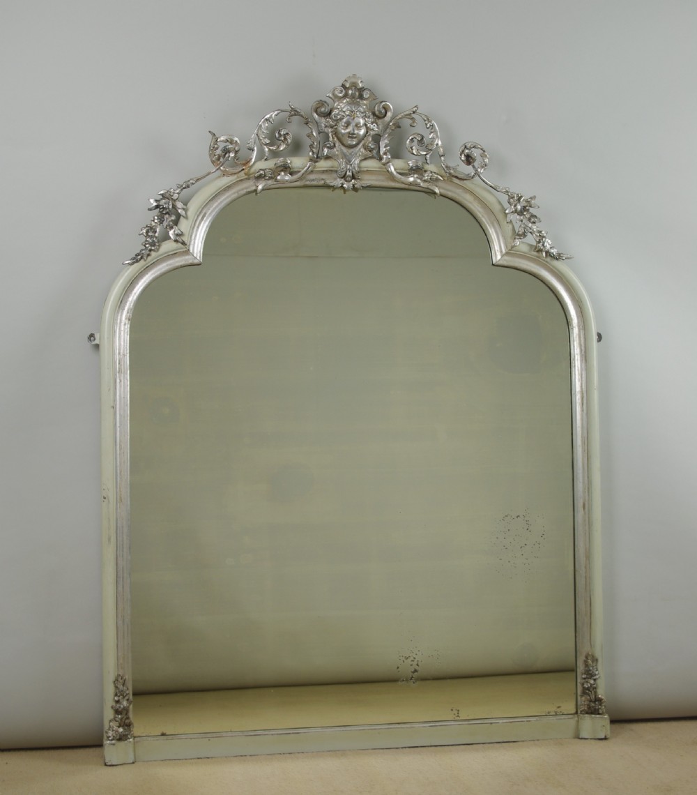 An arched Victorian overmantel mirror featuring a cream painted frame with silvered detailing and an ornate carved mask cresting.