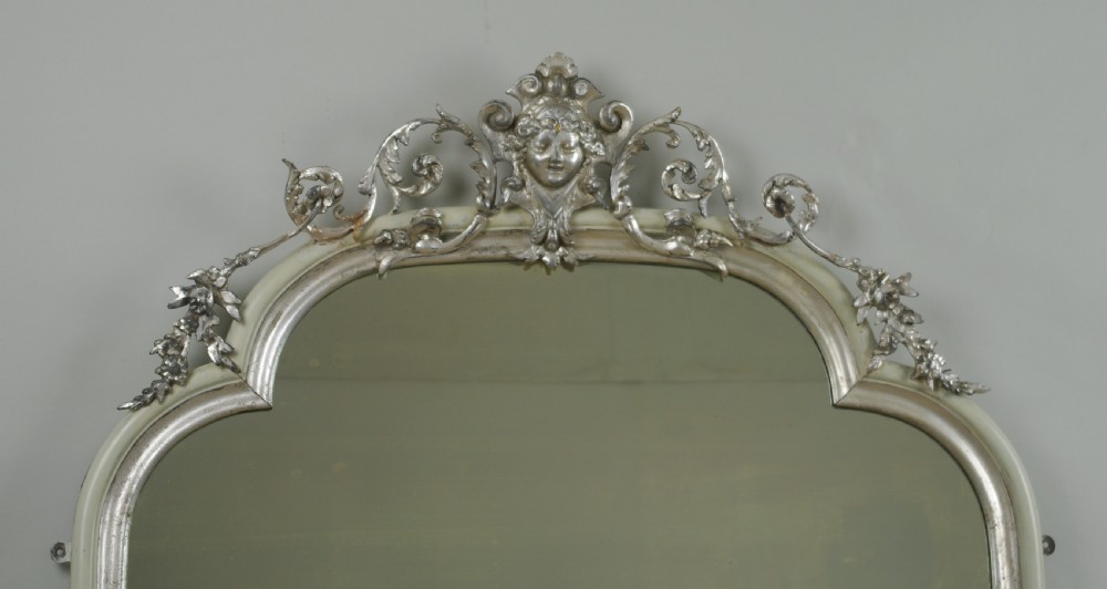 An arched Victorian overmantel mirror featuring a cream painted frame with silvered detailing and an ornate carved mask cresting. View II