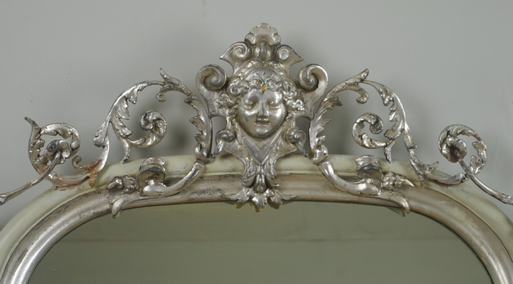 An arched Victorian overmantel mirror featuring a cream painted frame with silvered detailing and an ornate carved mask cresting. View III