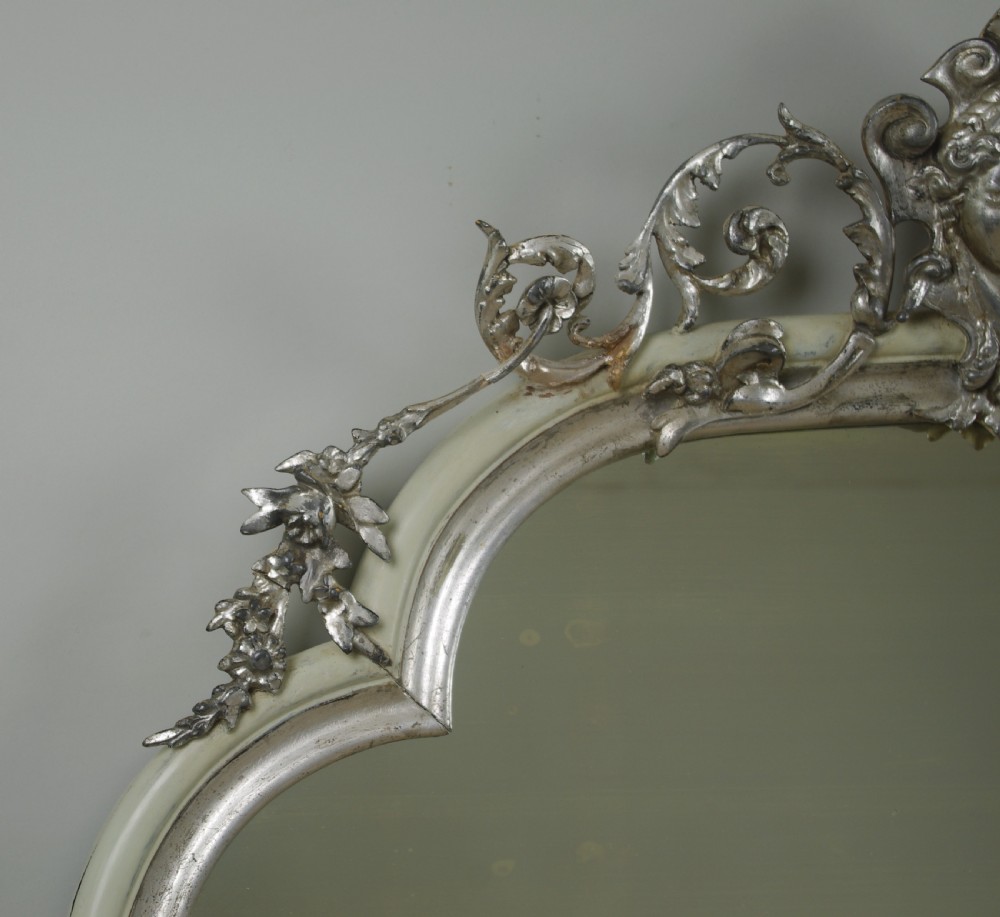 An arched Victorian overmantel mirror featuring a cream painted frame with silvered detailing and an ornate carved mask cresting. View IV
