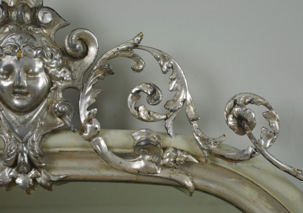 An arched Victorian overmantel mirror featuring a cream painted frame with silvered detailing and an ornate carved mask cresting. View VI
