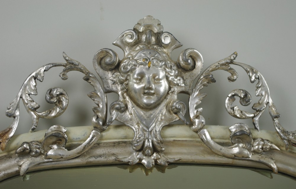 An arched Victorian overmantel mirror featuring a cream painted frame with silvered detailing and an ornate carved mask cresting. View VII