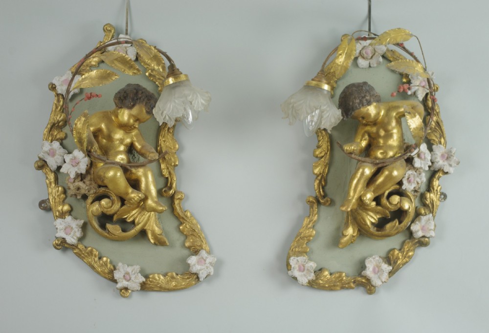 A pair of 19th-century French gilt wall lights featuring sculpted putti figures seated within asymmetrical gilded Rococo floral frames with frosted glass flower shades.