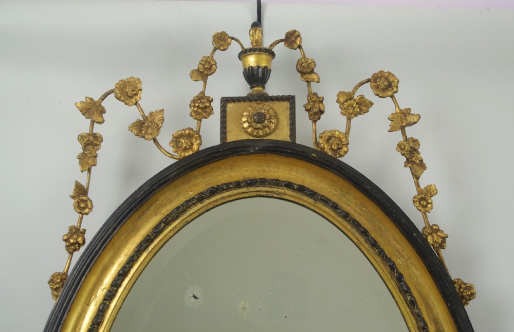 A Victorian oval wall mirror featuring an ebonised wood frame with a parcel-gilt inner border, an urn cresting, and floral swag decorations. View II