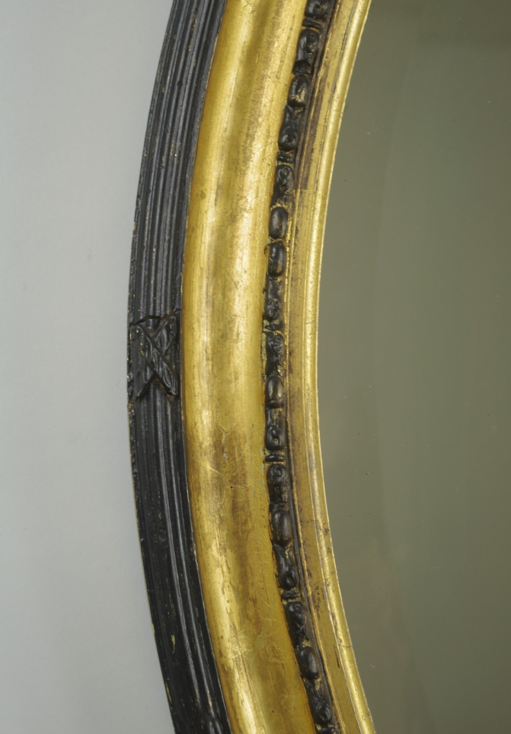 A Victorian oval wall mirror featuring an ebonised wood frame with a parcel-gilt inner border, an urn cresting, and floral swag decorations. View V
