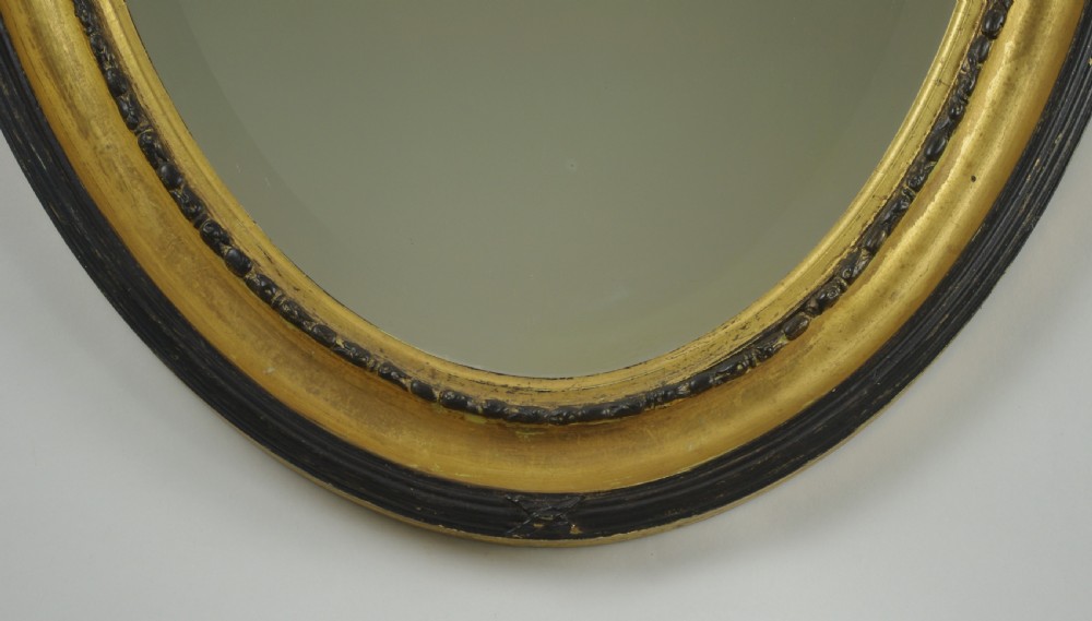 A Victorian oval wall mirror featuring an ebonised wood frame with a parcel-gilt inner border, an urn cresting, and floral swag decorations. View VI