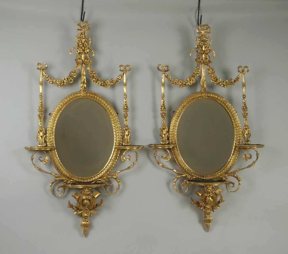 A pair of gilded oval wall mirrors featuring top ribbon bow cresting, hanging floral garlands, and small integrated display shelves.