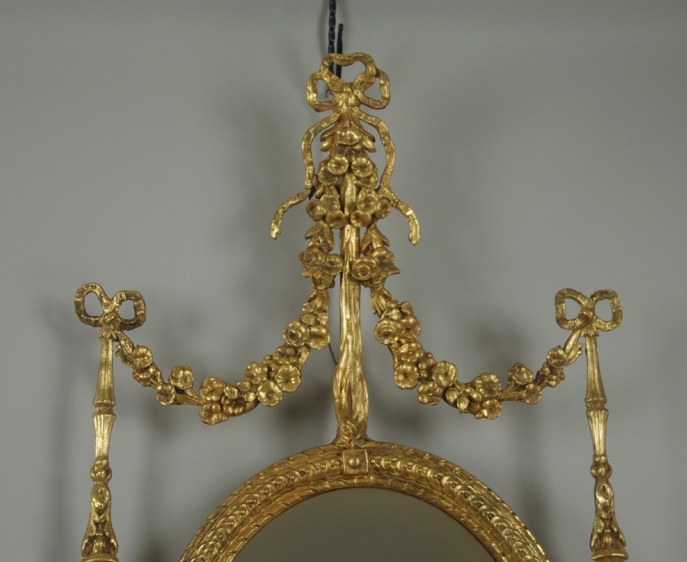 A pair of gilded oval wall mirrors featuring top ribbon bow cresting, hanging floral garlands, and small integrated display shelves. View V