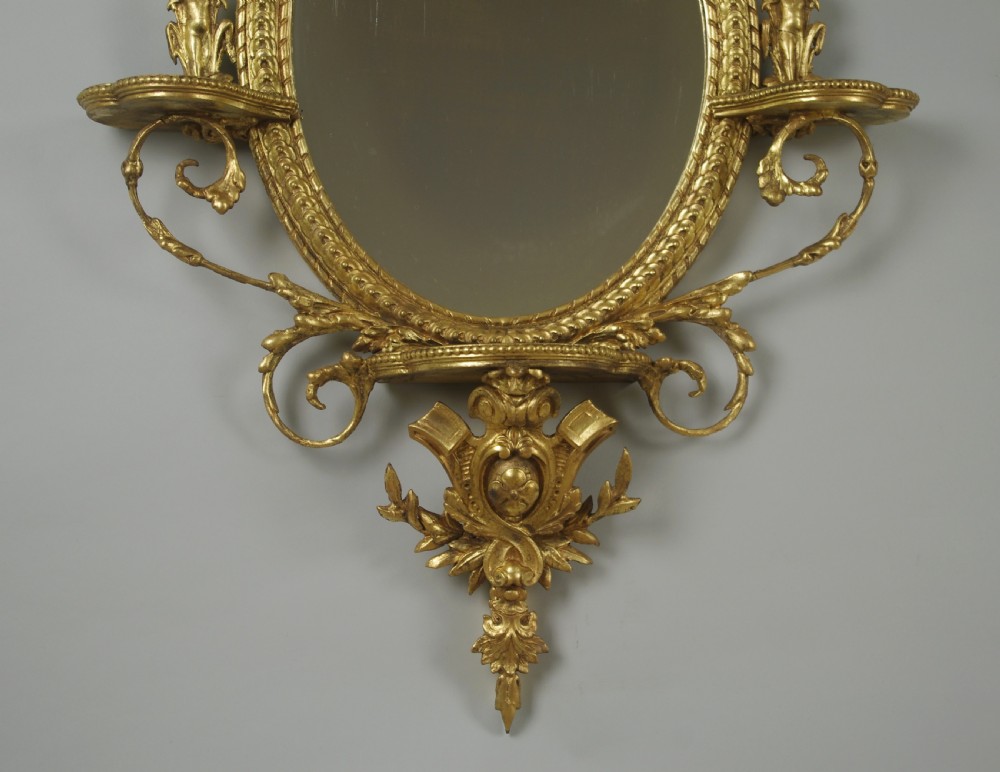 A pair of gilded oval wall mirrors featuring top ribbon bow cresting, hanging floral garlands, and small integrated display shelves. View VI