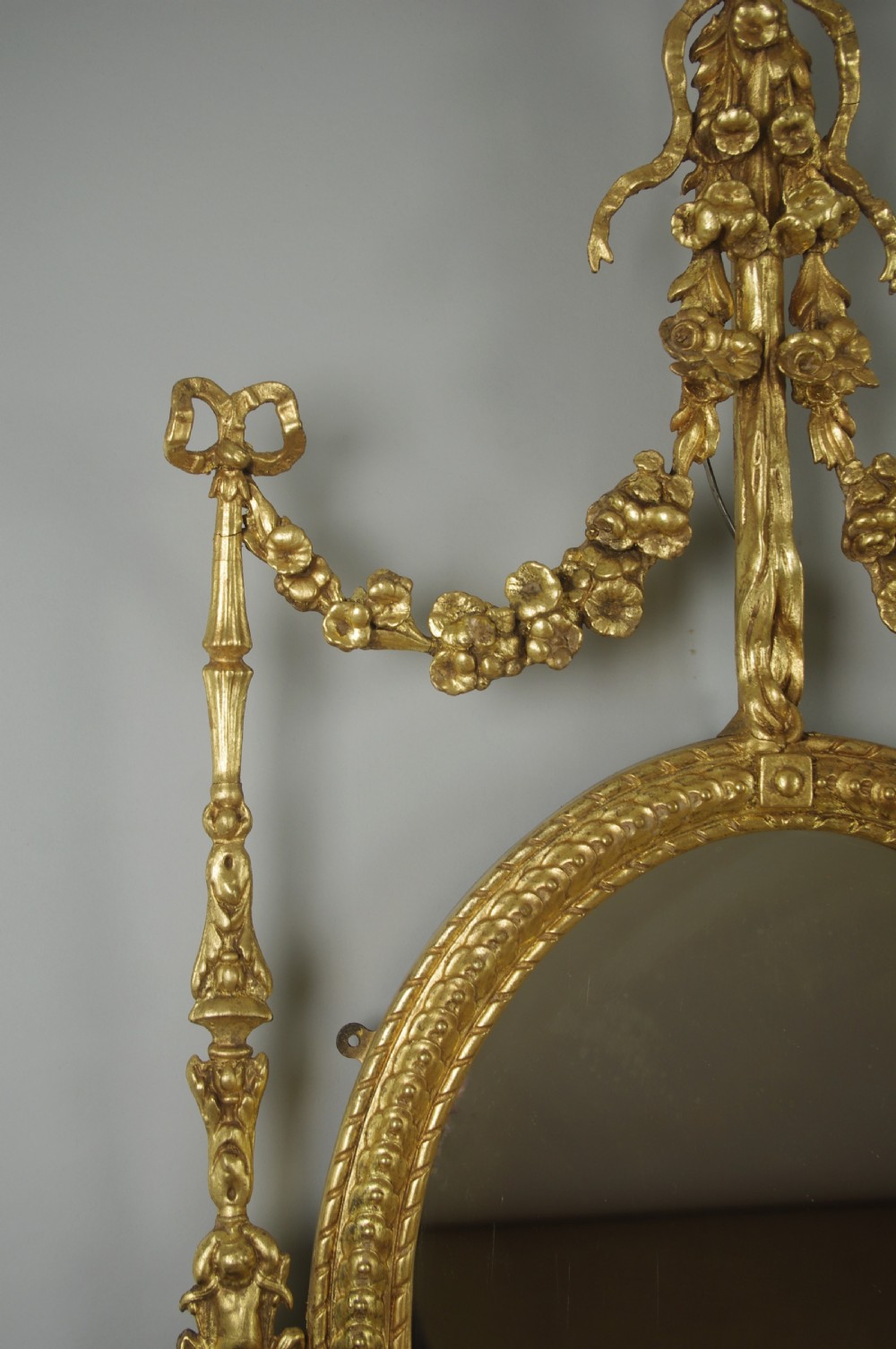 A pair of gilded oval wall mirrors featuring top ribbon bow cresting, hanging floral garlands, and small integrated display shelves. View VIII