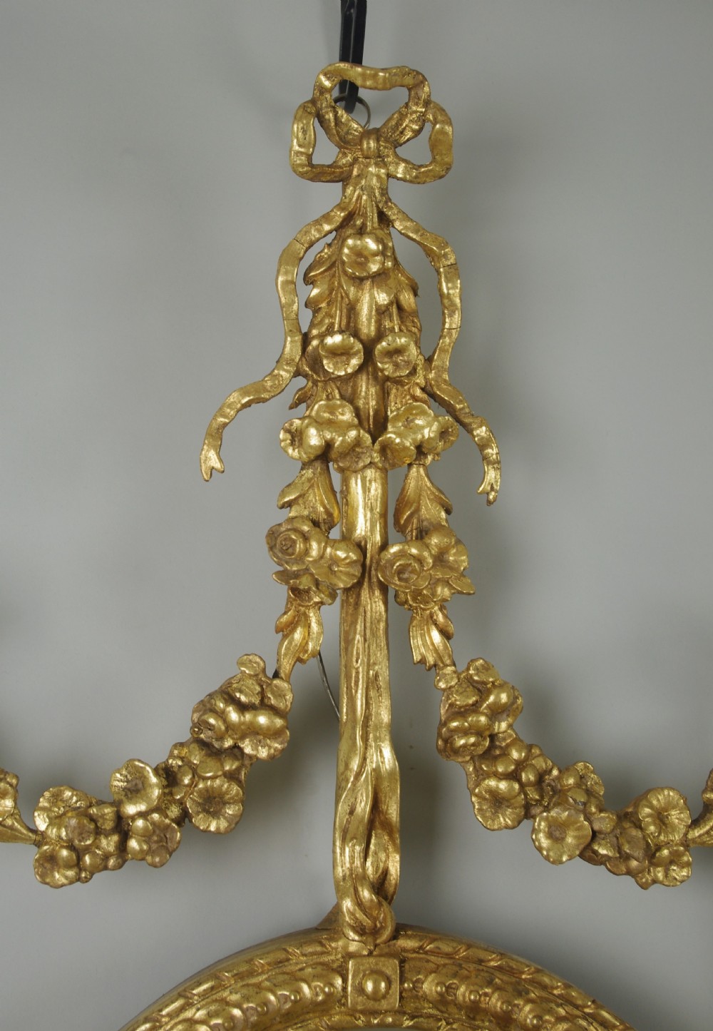 A pair of gilded oval wall mirrors featuring top ribbon bow cresting, hanging floral garlands, and small integrated display shelves. View IX