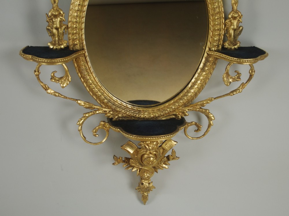 A pair of gilded oval wall mirrors featuring top ribbon bow cresting, hanging floral garlands, and small integrated display shelves. View XII
