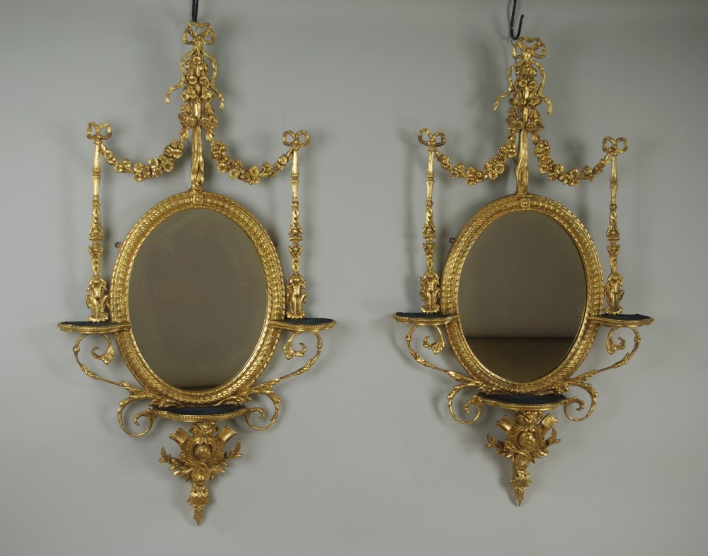 A pair of gilded oval wall mirrors featuring top ribbon bow cresting, hanging floral garlands, and small integrated display shelves. View XIII