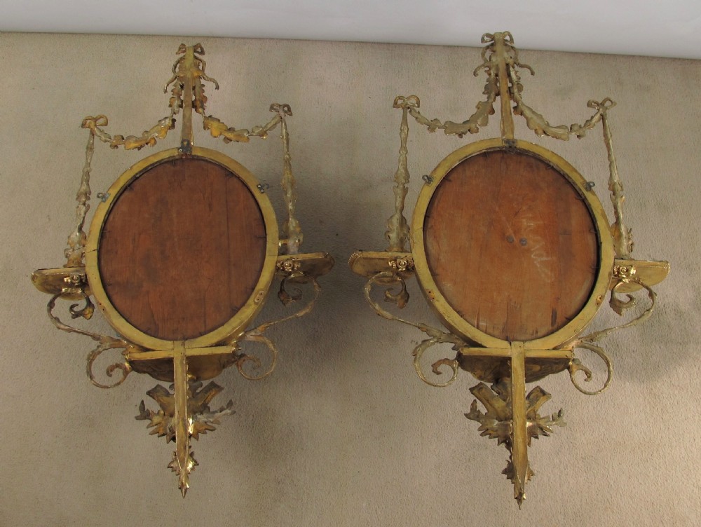A pair of gilded oval wall mirrors featuring top ribbon bow cresting, hanging floral garlands, and small integrated display shelves. View XIV