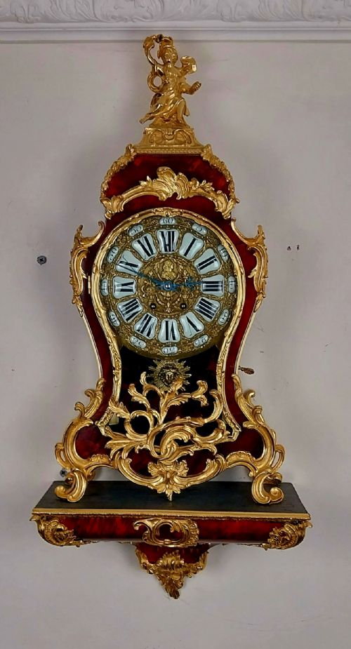 fig 1. An antique French Rococo revival Boulle wall clock featuring a red tortoiseshell case adorned with elaborate gilt bronze mounts, a figure finial, and an enamel cartouche dial on a matching wall bracket.
