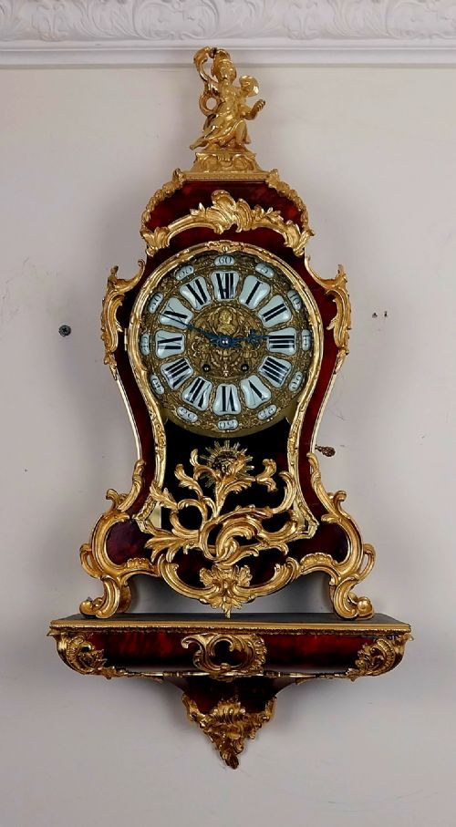 fig 2. An antique French Rococo revival Boulle wall clock featuring a red tortoiseshell case adorned with elaborate gilt bronze mounts, a figure finial, and an enamel cartouche dial on a matching wall bracket.
