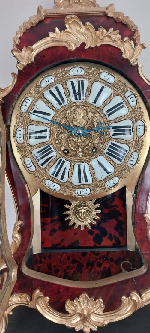 fig 3. An antique French Rococo revival Boulle wall clock featuring a red tortoiseshell case adorned with elaborate gilt bronze mounts, a figure finial, and an enamel cartouche dial on a matching wall bracket.