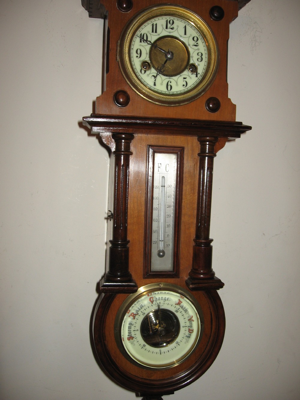 A late Victorian carved wooden wall clock featuring an upper clock face, a central thermometer, and a lower barometer dial. View V