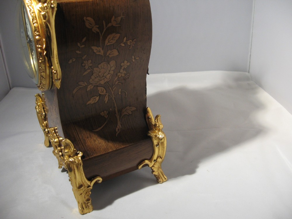 A nineteenth-century French Rococo style mantel clock crafted from dark rosewood with floral marquetry inlay, ornate gilt-bronze mounts, and a white enamel dial. View VIII