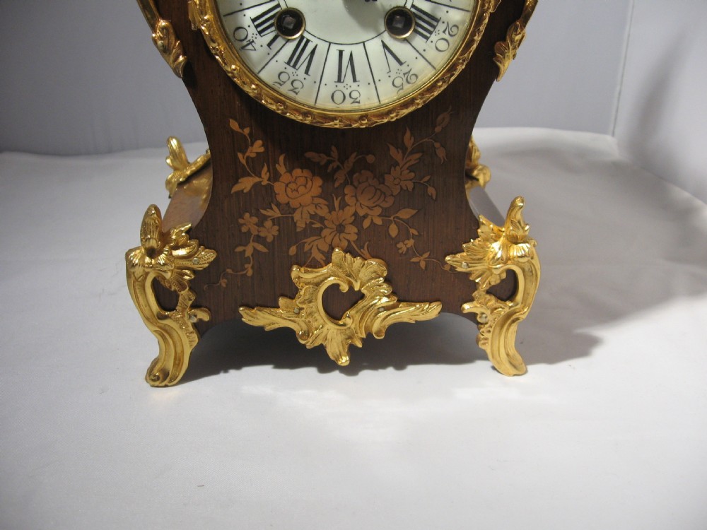 A nineteenth-century French Rococo style mantel clock crafted from dark rosewood with floral marquetry inlay, ornate gilt-bronze mounts, and a white enamel dial. View IX