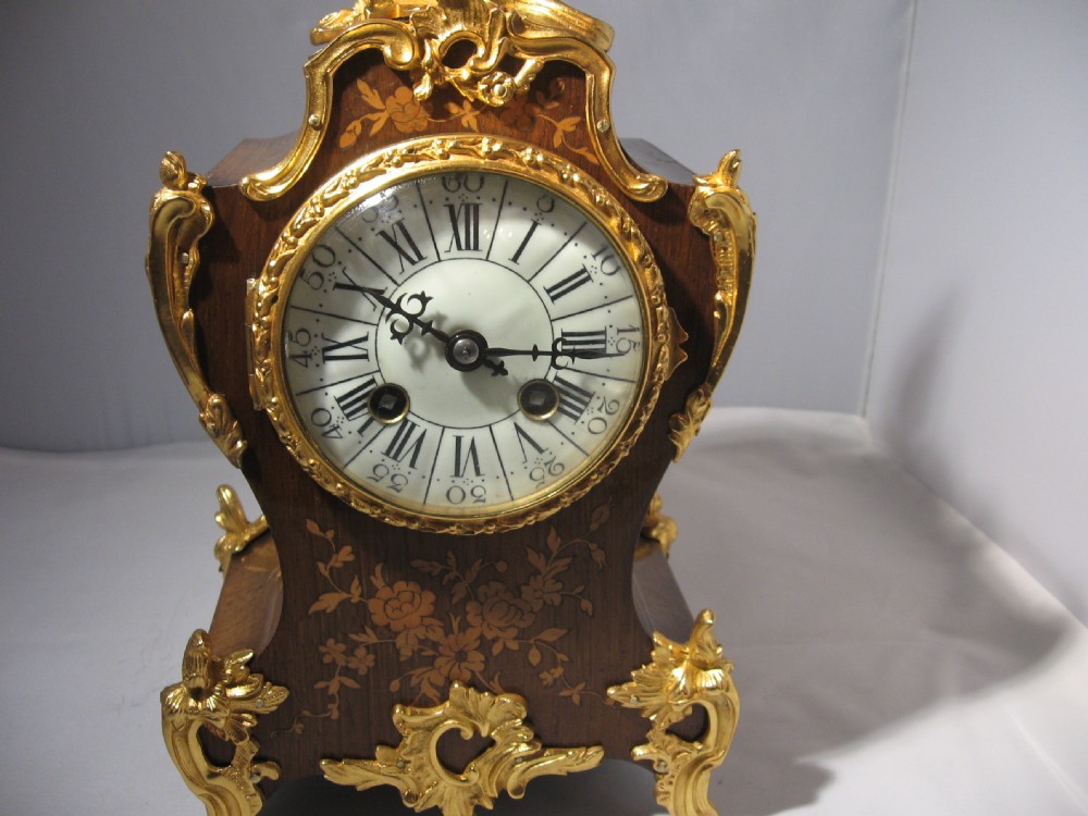 A nineteenth-century French Rococo style mantel clock crafted from dark rosewood with floral marquetry inlay, ornate gilt-bronze mounts, and a white enamel dial. View X