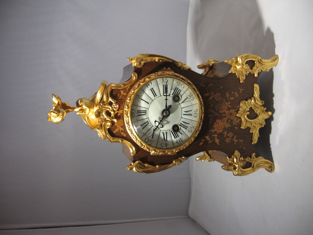 A nineteenth-century French Rococo style mantel clock crafted from dark rosewood with floral marquetry inlay, ornate gilt-bronze mounts, and a white enamel dial. View XII