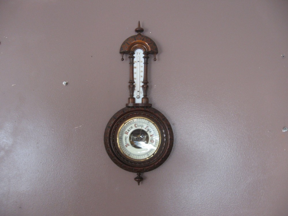 A carved dark walnut wall barometer featuring a vertical thermometer at the top and a circular aneroid pressure dial at the base.
