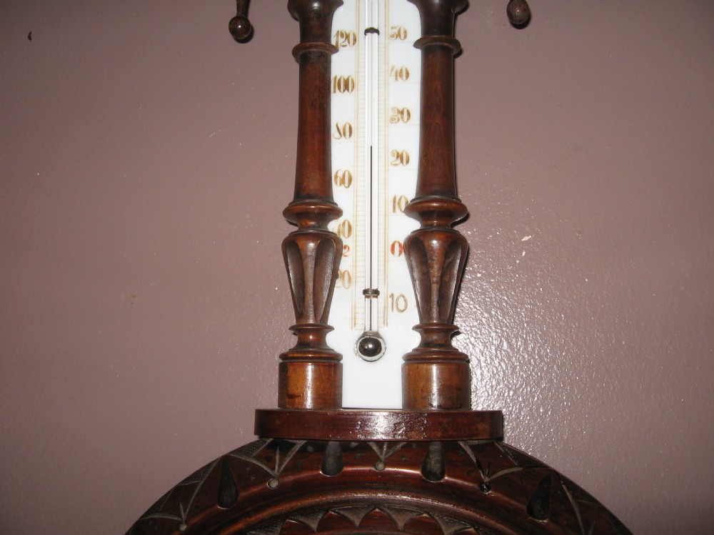 A carved dark walnut wall barometer featuring a vertical thermometer at the top and a circular aneroid pressure dial at the base. View V