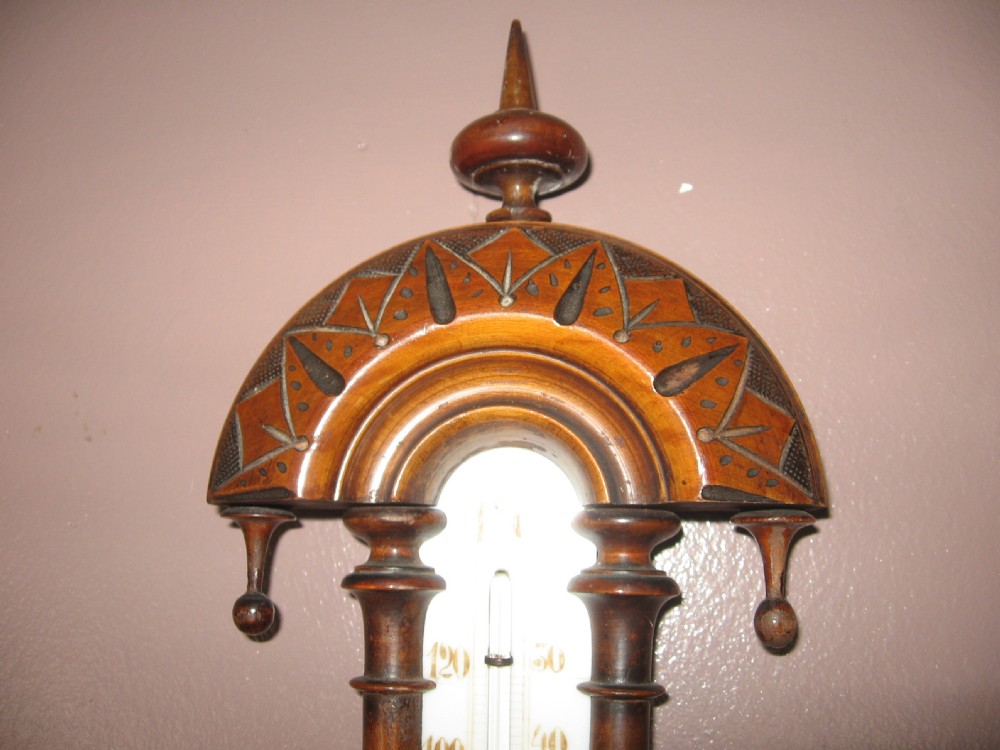 A carved dark walnut wall barometer featuring a vertical thermometer at the top and a circular aneroid pressure dial at the base. View VI