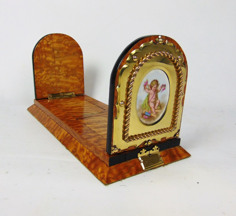An expanding wooden book slide featuring golden burr wood veneer, folding arched ends, and a gilt brass front plate framing an oval painted porcelain cherub medallion.