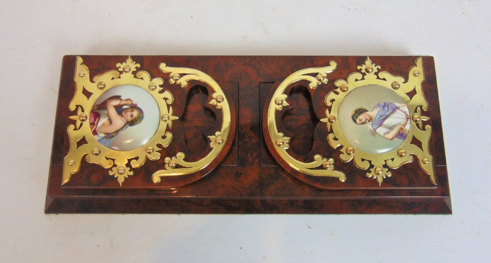 An antique burr walnut book slide featuring pierced brass strapwork with cut-out motifs and two inset circular porcelain plaques painted with female portraits.