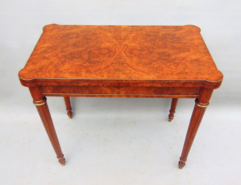 A Victorian burr walnut fold-over card table featuring a figured veneer top, brass beaded edging, and turned fluted legs.