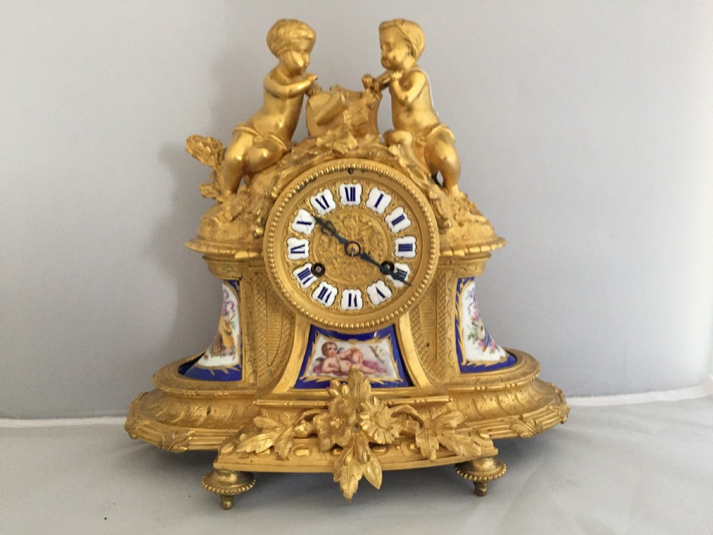 An antique French gilt bronze ormolu mantel clock featuring sculpted cherubs on top and hand-painted blue porcelain panels around a Roman numeral dial.
