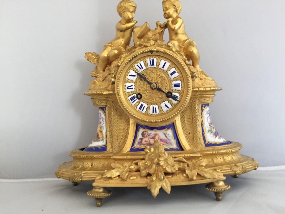 An antique French gilt bronze ormolu mantel clock featuring sculpted cherubs on top and hand-painted blue porcelain panels around a Roman numeral dial. View II