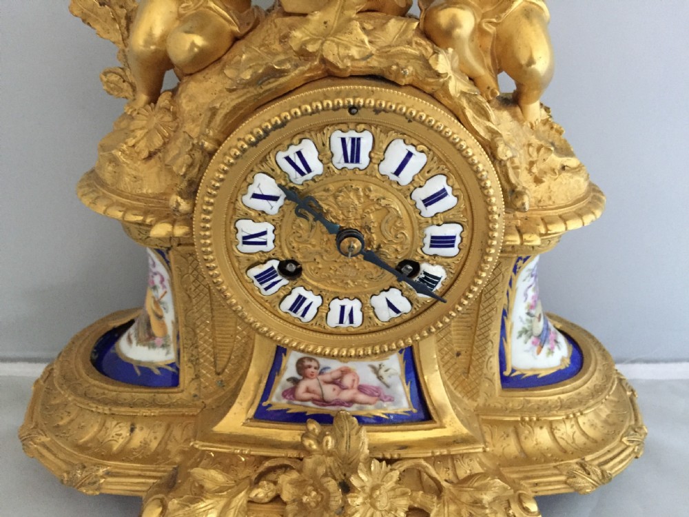 An antique French gilt bronze ormolu mantel clock featuring sculpted cherubs on top and hand-painted blue porcelain panels around a Roman numeral dial. View V
