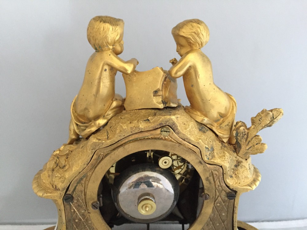 An antique French gilt bronze ormolu mantel clock featuring sculpted cherubs on top and hand-painted blue porcelain panels around a Roman numeral dial. View IX